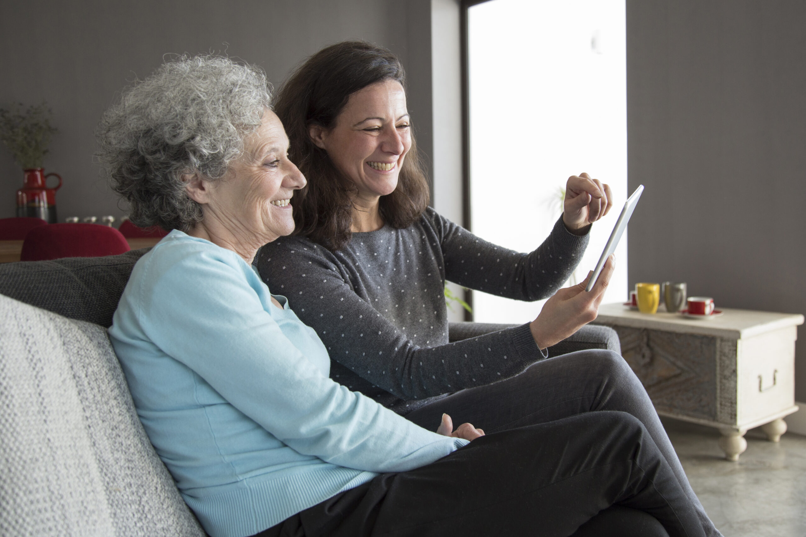 Happy elderly woman and her daughter browsing on tablet computer. Mother and daughter sitting on couch with home interior in background. Technology concept.